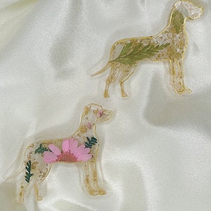 May include: Two resin dog ornaments featuring pressed flowers and gold leaf. The dog on the left has a pink flower, while the dog on the right has green leaves.