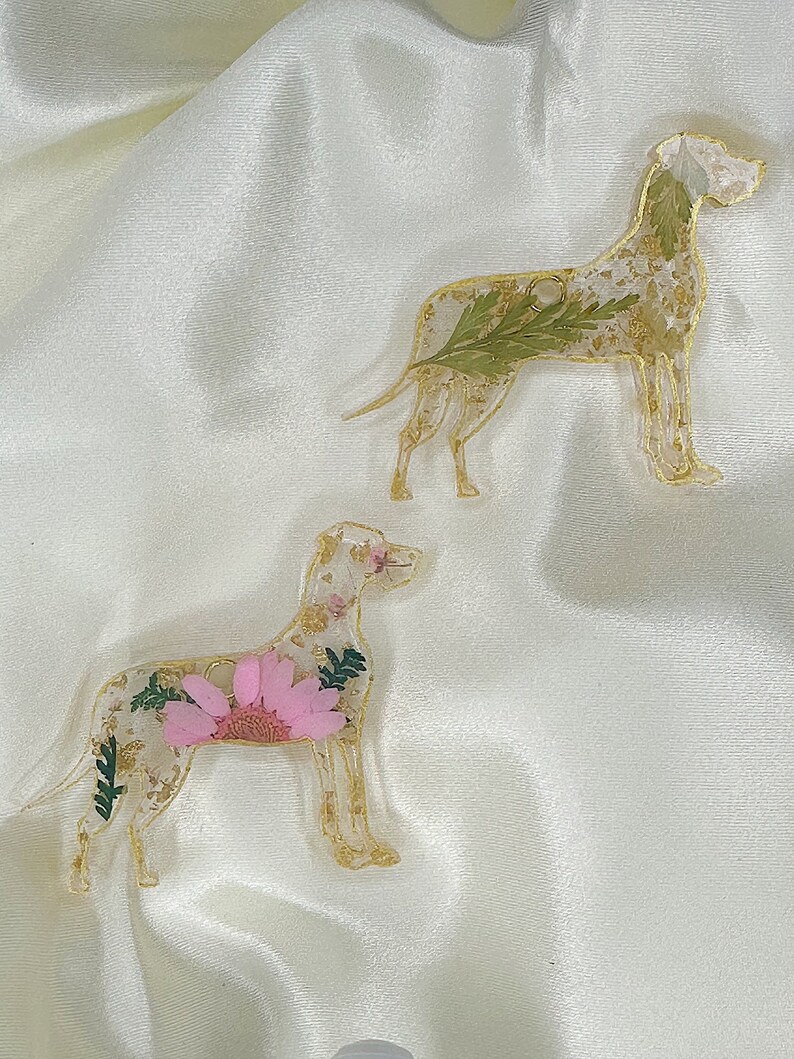 May include: Two resin dog ornaments featuring pressed flowers and gold leaf. The dog on the left has a pink flower, while the dog on the right has green leaves.