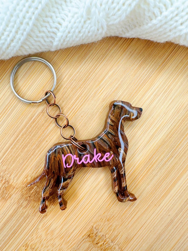 May include: A brown and black resin dog keychain with the name "Drake" written in pink lettering. The keychain is attached to a silver key ring.