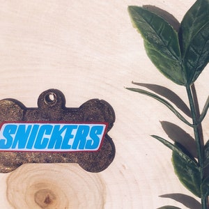 May include: A brown bone-shaped dog tag with the word "SNICKERS" printed in blue letters on a red background.