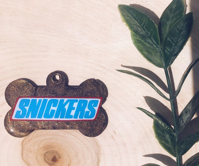 May include: A brown bone-shaped dog tag with the word "SNICKERS" printed in blue letters on a red background.