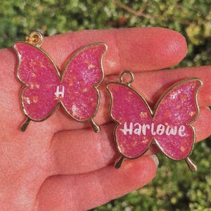 Puede incluir: Dos encantos de mariposa rosa con detalles dorados. Un encanto tiene la letra "H" y el otro encanto tiene el nombre "Harlowe".