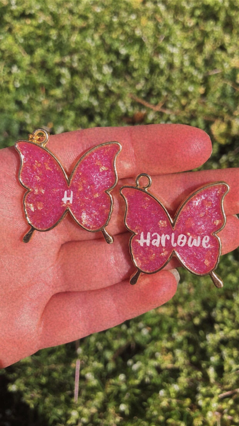 Puede incluir: Dos encantos de mariposa rosa con detalles dorados. Un encanto tiene la letra "H" y el otro encanto tiene el nombre "Harlowe".