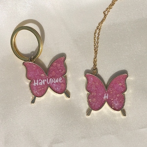 Puede incluir: Dos llaveros de mariposa dorados con purpurina rosa. Un llavero tiene el nombre "Harlowe" escrito en &eacute;l, y el otro tiene la letra "H" escrita en &eacute;l.