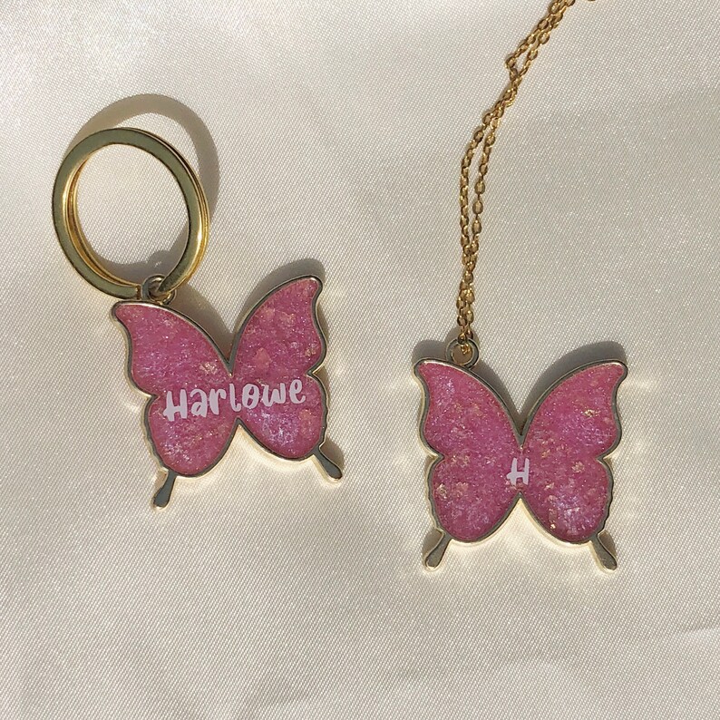 Puede incluir: Dos llaveros de mariposa dorados con purpurina rosa. Un llavero tiene el nombre "Harlowe" escrito en &eacute;l, y el otro tiene la letra "H" escrita en &eacute;l.