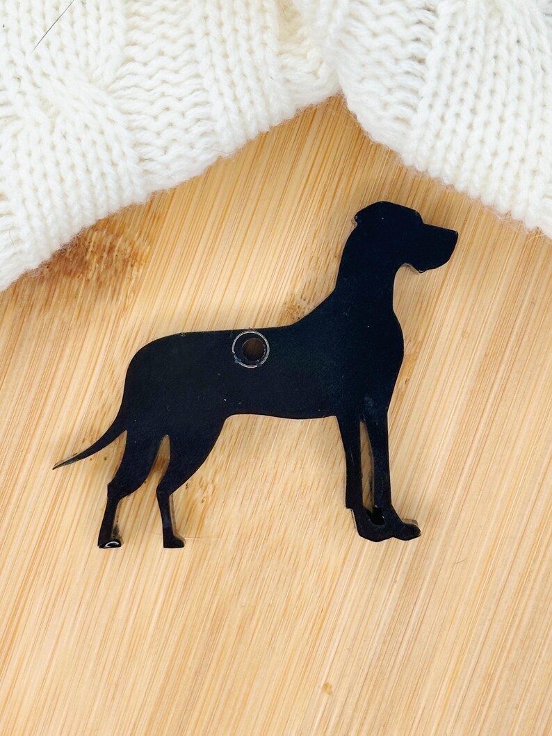 May include: Black silhouette of a Great Dane dog, cut from wood, with a hole for hanging. The silhouette is on a light brown wooden surface.