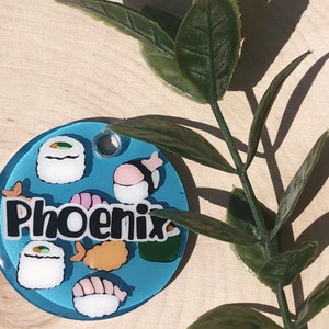 May include: A blue and white round tag with the word "Phoenix" in black lettering. The tag is decorated with images of sushi rolls, shrimp, and seaweed.
