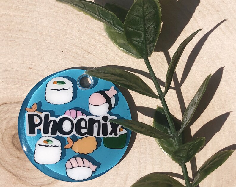 May include: A blue and white round tag with the word "Phoenix" in black lettering. The tag is decorated with images of sushi rolls, shrimp, and seaweed.