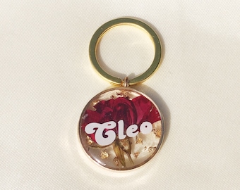May include: Gold key chain with a round resin charm featuring a pressed red flower and gold leaf accents. The charm has the name "Cleo" written in white cursive.