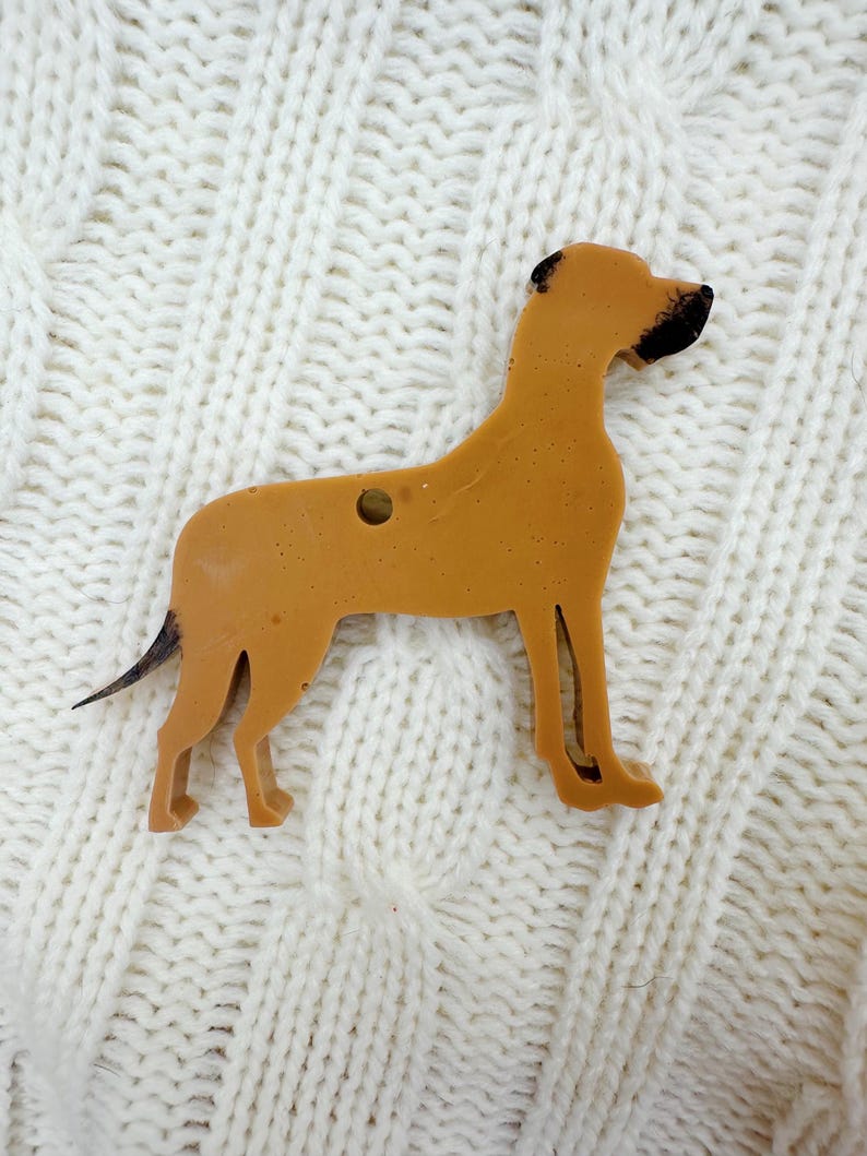 May include: A brown wooden cutout of a dog, possibly a Great Dane, with black markings on its head. The cutout is on a white knitted surface.