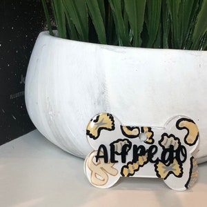 May include: White bone-shaped dog tag with a black and white pasta pattern and the name "Alfredo" written in black.