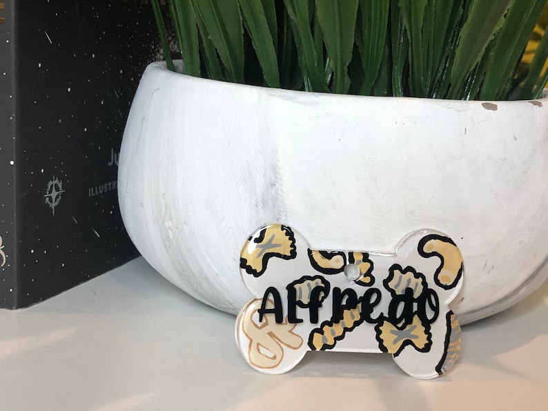 May include: White bone-shaped dog tag with a black and white pasta pattern and the name "Alfredo" written in black.