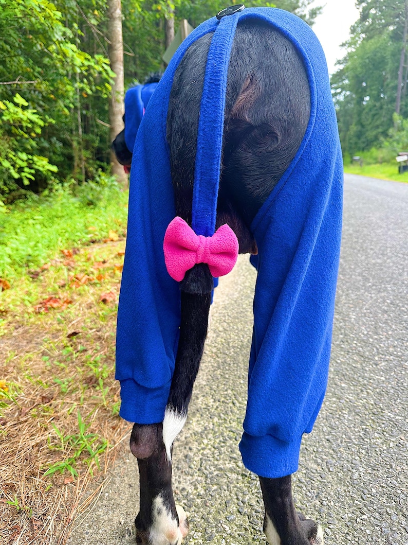 May include: A royal blue fleece dog jumpsuit featuring long sleeves with ribbed cuffs, a black button closure at the top, and a bright pink fabric bow attached to the tail opening. The garment is designed to fit around the tail and hind legs.
