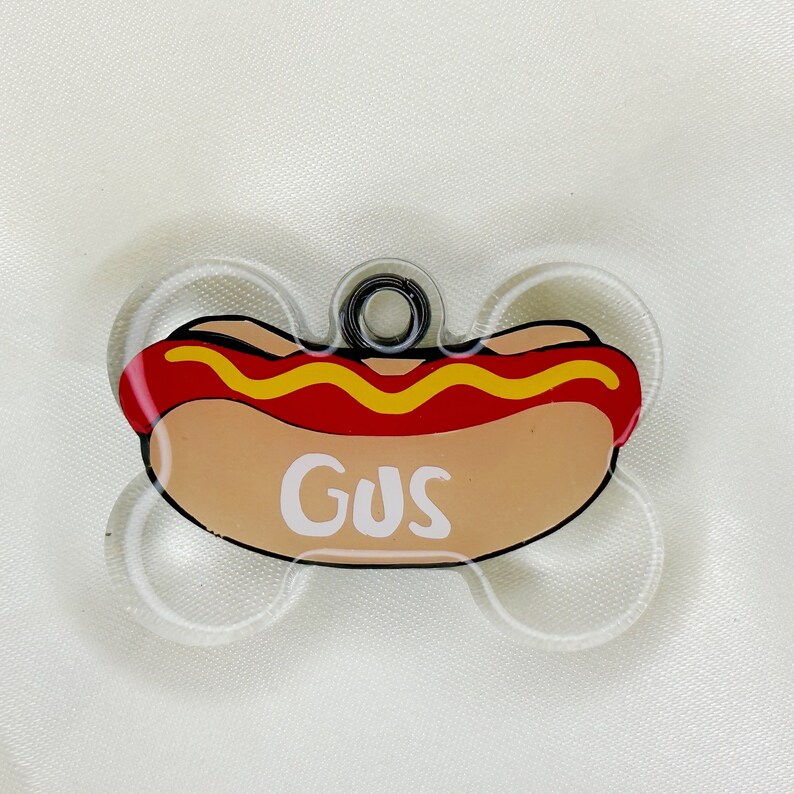 May include: A clear plastic dog tag shaped like a bone with a red and yellow hot dog design and the name "Gus" printed in white.