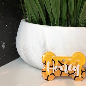 May include: A yellow bone-shaped dog tag with a honeycomb pattern and the name "Honey" written in white lettering.