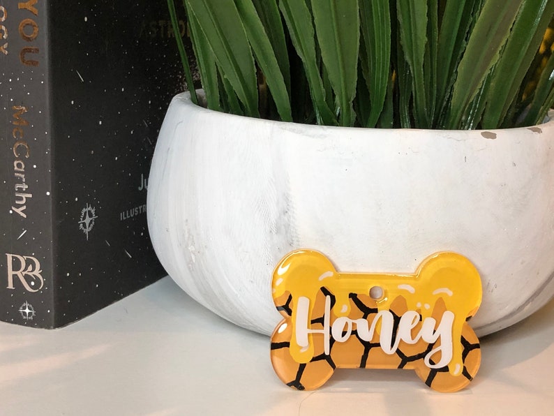 May include: A yellow bone-shaped dog tag with a honeycomb pattern and the name "Honey" written in white lettering.