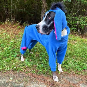 May include: A blue fleece dog jumpsuit featuring a hood with long, floppy ears lined in pink, a black tuft of hair on top, and a small pink bow attached to the rear. The garment covers the torso and legs, providing a full-body fit for the animal.