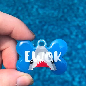 Puede incluir: Chapa para perro de hueso azul con un gr&aacute;fico de tibur&oacute;n blanco y el nombre "EWOK" en texto blanco.