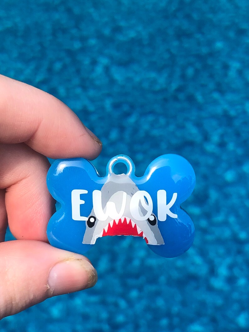 Puede incluir: Chapa para perro de hueso azul con un gr&aacute;fico de tibur&oacute;n blanco y el nombre "EWOK" en texto blanco.