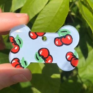 May include: A white bone-shaped dog tag with a cherry pattern. The tag is made of acrylic and has a hole for a leash.