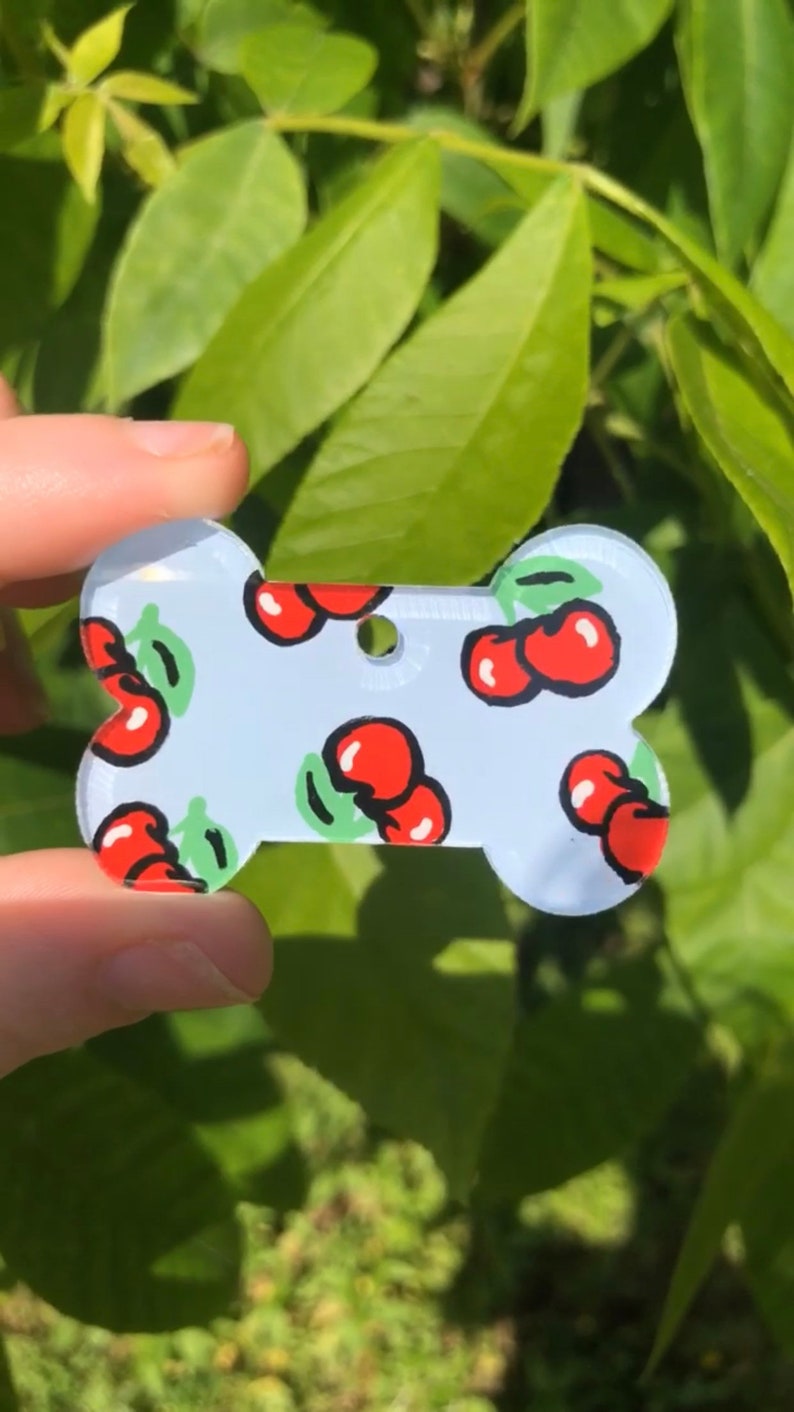 May include: A white bone-shaped dog tag with a cherry pattern. The tag is made of acrylic and has a hole for a leash.