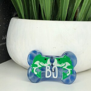 Puede incluir: Una etiqueta para perro en forma de hueso azul y blanco con un dise&ntilde;o de pez verde y el nombre "BO" impreso en ella.