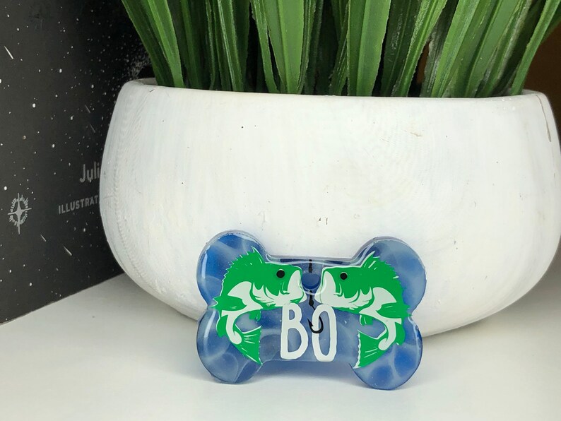 Puede incluir: Una etiqueta para perro en forma de hueso azul y blanco con un dise&ntilde;o de pez verde y el nombre "BO" impreso en ella.