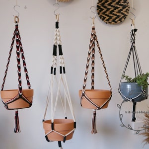 Puede incluir: Tres maceteros colgantes de macramé con cordones y borlas de diferentes colores. Los maceteros sostienen macetas de terracota, una con una planta verde y dos vacías.