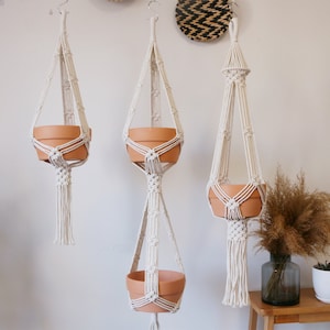 Op de afbeelding: Drie macramé plantenhangers met terracotta potten. De hangers zijn gemaakt van wit katoenen koord en hebben een eenvoudig, minimalistisch ontwerp. De potten worden door een reeks knopen aan de hangers opgehangen.