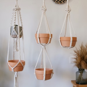 Puede incluir: Tres maceteros colgantes de macramé con macetas de terracota. Los maceteros están hechos de cordón de algodón blanco y tienen un diseño simple y minimalista. Las macetas cuelgan del techo y están llenas de plantas.
