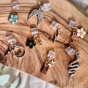May include: Assortment of charms for jewellery making, including a flower, wings, lightning bolt, snake, letter M with a bee, a key with a crescent moon, and a flower with a pink centre. The charms are on a wooden surface.