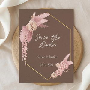 Puede incluir: Tarjeta Save the Date marrón con un marco geométrico dorado y detalles florales. La tarjeta presenta el texto "Save the Date", nombres y una fecha. La tarjeta está sobre una superficie de madera con una cinta blanca.