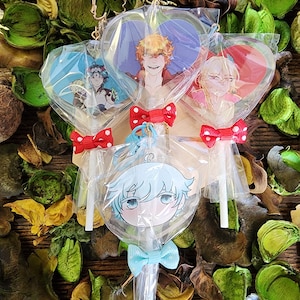 Puede incluir: Tres piruletas en forma de corazón con diferentes personajes de anime impresos en ellas. Las piruletas están envueltas en plástico transparente y tienen lazos de lunares rojos y blancos. Las piruletas están sobre una superficie de madera con hojas verdes y marrones.