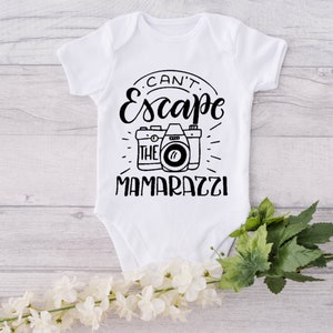 May include: White baby onesie with black text that reads "Can't Escape The Mamarazzi" with a black and white illustration of a camera.