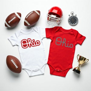 May include: Two white and red baby onesies with the word "Ohio" in red and gray lettering. The onesies are surrounded by footballs, a football helmet, and a stopwatch.