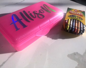 Op de afbeelding: Een roze plastic potlooddoos met een glinsterende afwerking. De doos heeft de naam "Allison" in zilveren letters op de bovenkant. Naast de doos ligt een doos met 24 Crayola-kleurpotloden.
