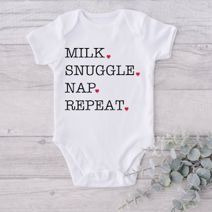 Op de afbeelding: Witte baby romper met zwarte tekst die "Milk, Snuggle, Nap, Repeat" luidt met een rood hartje na elk woord.