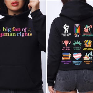 Op de afbeelding: Zwarte hoodie met de tekst "big fan of human rights" in kleurrijke letters op de voorkant. De achterkant heeft verschillende afbeeldingen en zinnen, waaronder "Black Lives Matter" en "Love is Love."