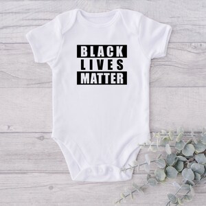 Può includere: Body bianco per neonato con testo nero in maiuscolo che recita "BLACK LIVES MATTER".
