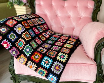 Puede incluir: Una colorida manta de ganchillo hecha a mano sobre un sillón de terciopelo rosa. La manta presenta un fondo negro con vibrantes patrones florales cuadrados de varios colores. El sillón tiene adornos verde oscuro.
