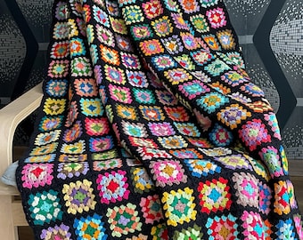 Könnte beinhalten: Eine handgefertigte Häkeldecke mit schwarzem Rand. Die Decke hat ein Patchwork-Design aus bunten Quadraten in verschiedenen Farbtönen, darunter Rot, Gelb, Blau und Grün. Die Decke ist über einen Holzstuhl drapiert.