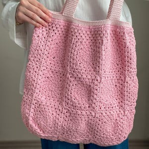 Puede incluir: Una bolsa de mano de crochet rosa con asas gruesas. La bolsa est&aacute; hecha con un delicado patr&oacute;n de puntadas y tiene una forma ligeramente redondeada.