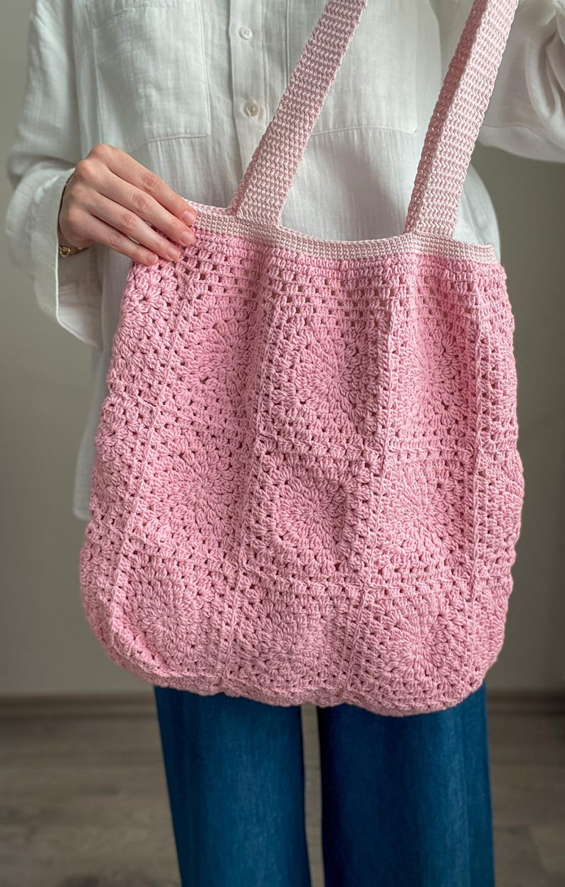 Puede incluir: Una bolsa de mano de crochet rosa con asas gruesas. La bolsa est&aacute; hecha con un delicado patr&oacute;n de puntadas y tiene una forma ligeramente redondeada.
