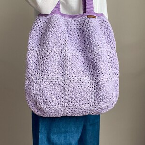 Puede incluir: Una bolsa de mano de crochet de color lavanda con una etiqueta de cuero marr&oacute;n. La bolsa la lleva una persona que lleva una camisa blanca y unos vaqueros azules.