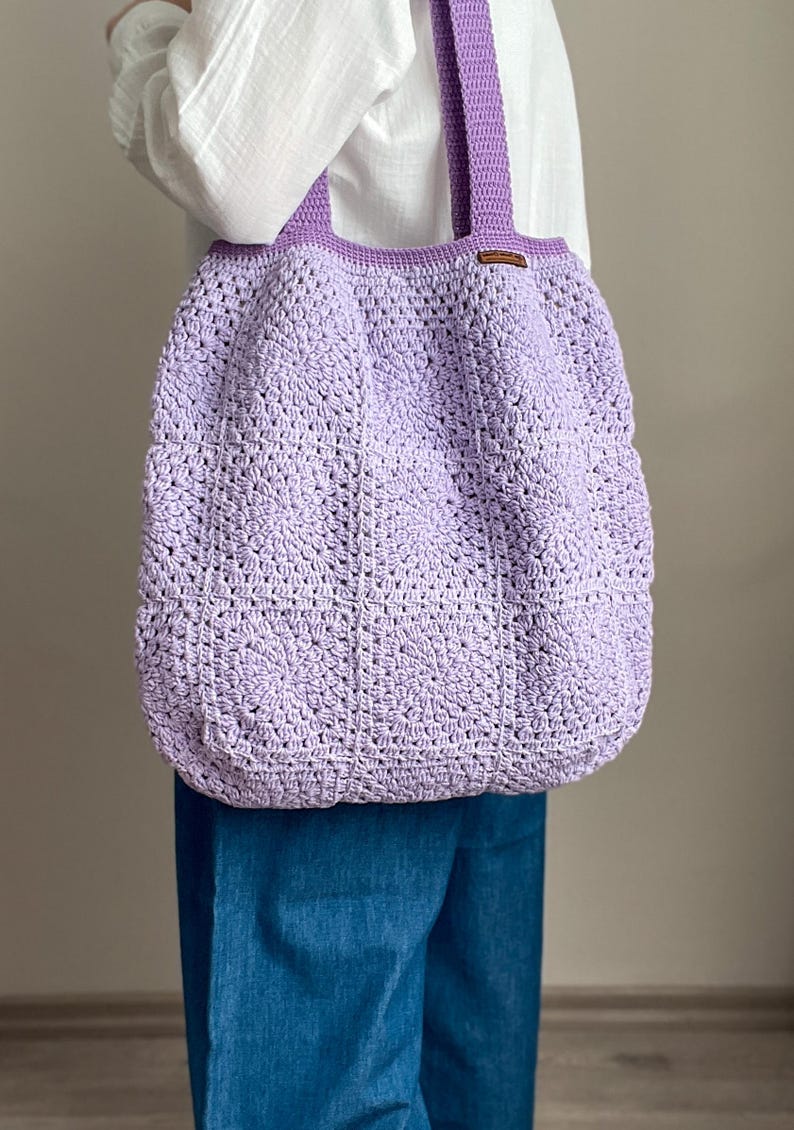 Puede incluir: Una bolsa de mano de crochet de color lavanda con una etiqueta de cuero marr&oacute;n. La bolsa la lleva una persona que lleva una camisa blanca y unos vaqueros azules.