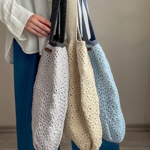 Puede incluir: Tres bolsas de mano de crochet en diferentes colores: gris claro, beige y azul claro. Las bolsas tienen una textura tejida y est&aacute;n colgadas del hombro de una persona.