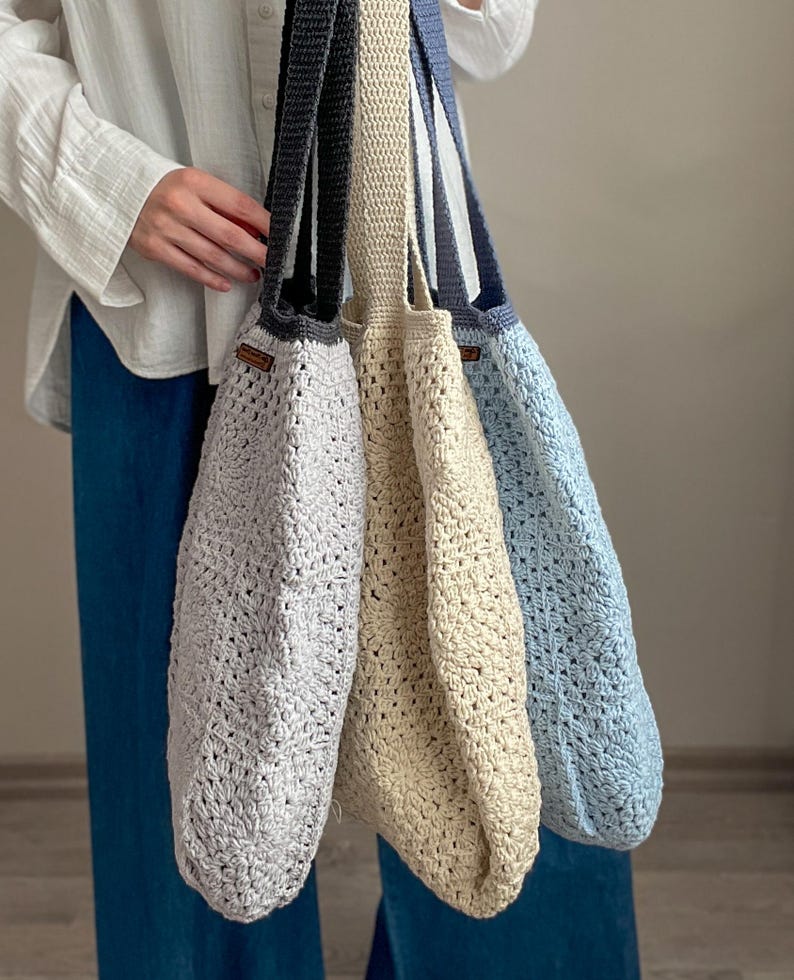 Puede incluir: Tres bolsas de mano de crochet en diferentes colores: gris claro, beige y azul claro. Las bolsas tienen una textura tejida y est&aacute;n colgadas del hombro de una persona.