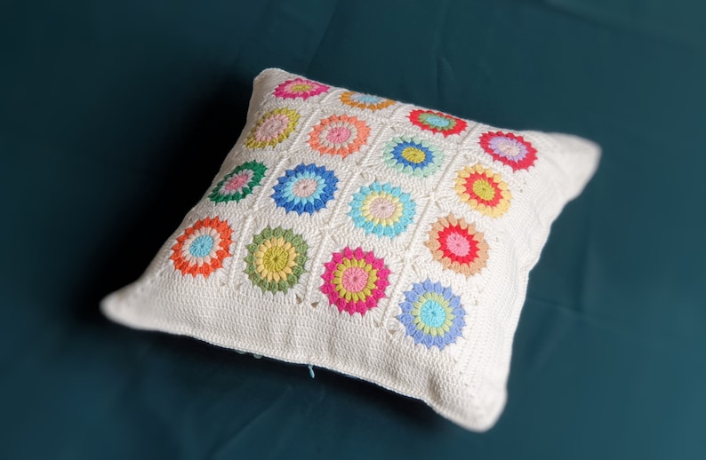 K&ouml;nnte beinhalten: Ein wei&szlig;es H&auml;kelkissen mit einem bunten Blumenmuster. Das Kissen ist aus einem weichen, strukturierten Stoff gefertigt und zeigt eine Vielzahl von Farben, darunter Rosa, Blau, Gr&uuml;n, Gelb und Orange.