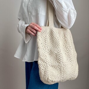 Puede incluir: Una bolsa de mano de crochet de color crema con una base ancha y plana y asas gruesas. La bolsa la lleva una persona que lleva una camisa blanca y pantalones vaqueros azules.