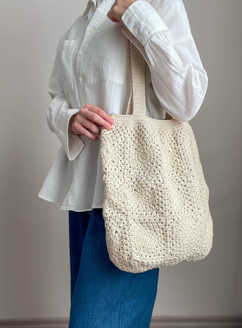 Puede incluir: Una bolsa de mano de crochet de color crema con una base ancha y plana y asas gruesas. La bolsa la lleva una persona que lleva una camisa blanca y pantalones vaqueros azules.